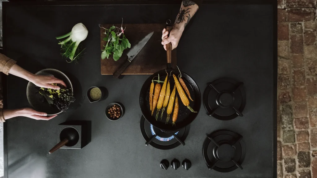 Top-down view of hands cooking on a PITT Cooking Cima 3 burner gas cooktop over a black kitchen bench top.